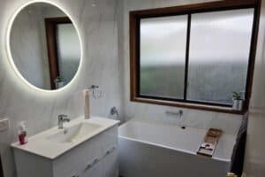 Bright bathroom with a round LED lit mirror, marble look tiled walls and a freestanding bath
